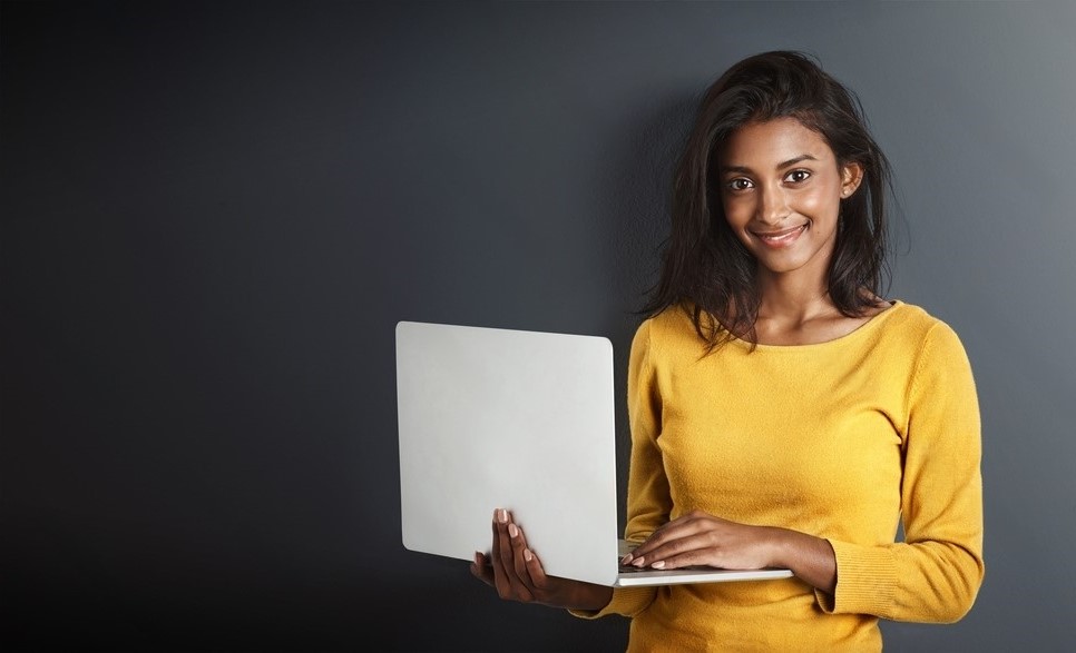 A woman in a yellow sweater stands against a dark gray background, holding an open laptop in her arms. She smiles softly at the camera, exuding a sense of warmth and familiarity that feels just like home.