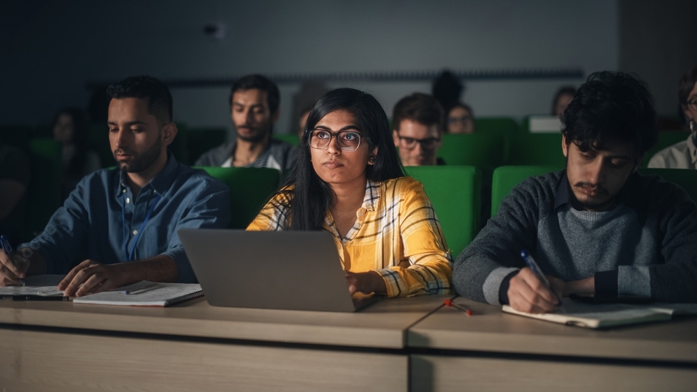 A group of students sits attentively in a lecture hall. One student in the middle uses a laptop, while others take notes on paper.