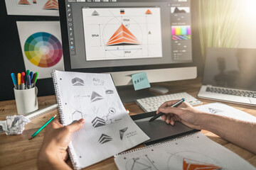 A designer’s hand sketching logos in a notebook with data charts on the computer screen in the background.